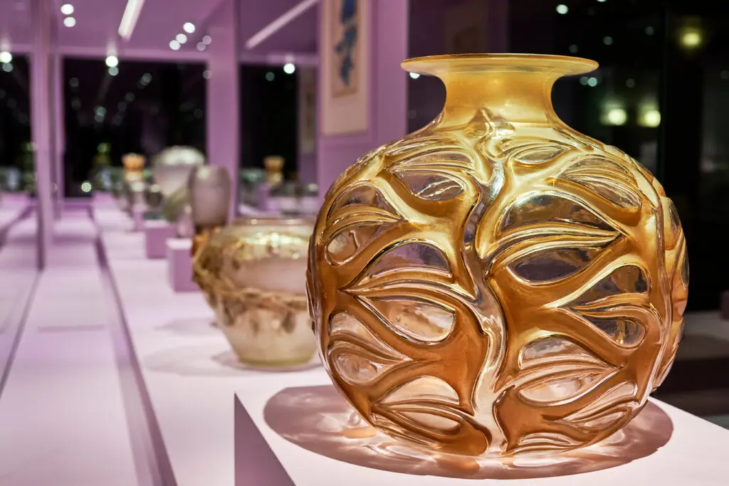 Переливающаяся желтая стеклянная ваза украшена узором виноградной лозы с прозрачными листьями. Он находится в галерее среди других стеклянных предметов.
